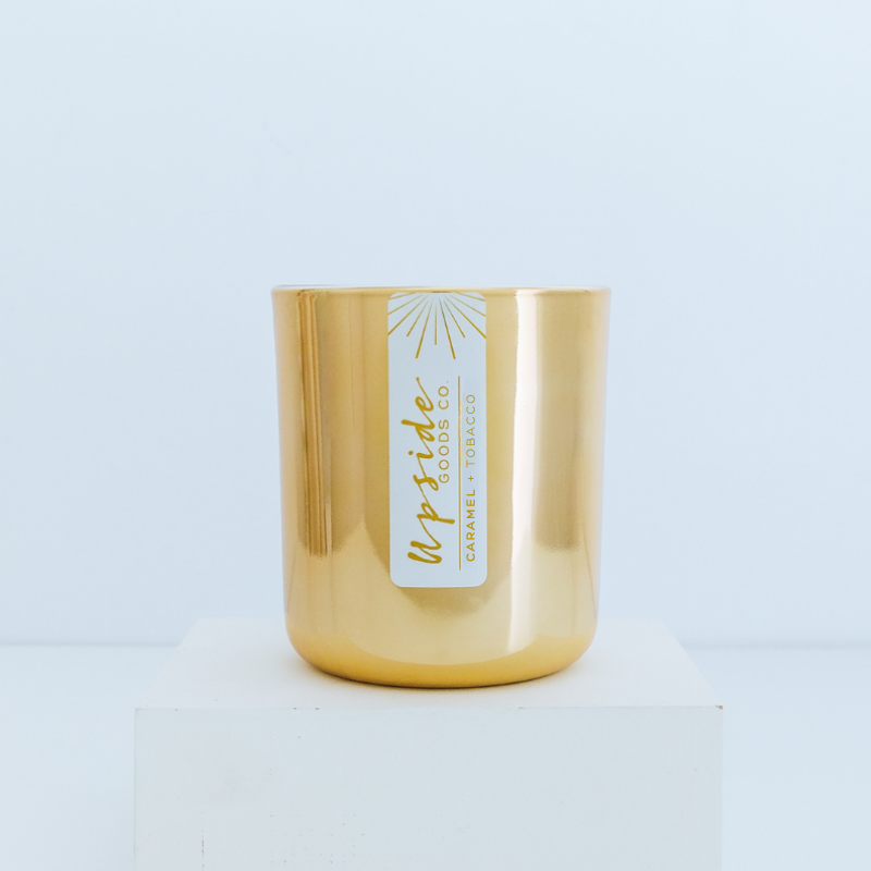 Gold candle with a label on a light gray background and Caramel + Tobacco Limited Edition scent. 